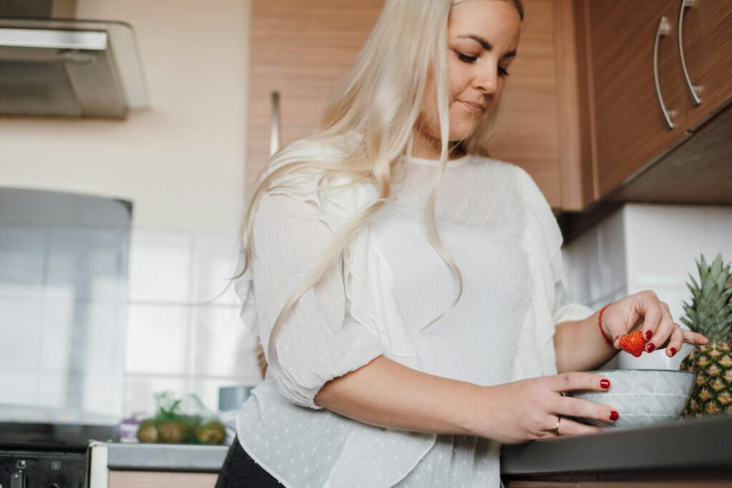 From below of crop concentrated housewife in casual wear standing in kitchen and adding strawberry into fruit salad while cooking in kitchen